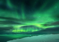 Aurora borealis over ocean. Northern lights in Teriberka, Russia. Starry sky with polar lights and clouds. Night winter landscape with aurora, sea with blurred water, snowy hills, stars. Travel