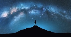 Milky Way arch and man on the mountain peak at starry night. Silhouette of alone guy, blue sky with bright stars in summer. Galaxy. Space background. Landscape with arched milky way. Travel and nature