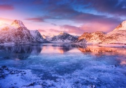 Aerial view of snowy mountains, blue sea with frosty coast, reflection in water, sky with pink clouds at colorful sunset. Lofoten islands, Norway. Winter landscape with snow covered rocks, fjord, ice