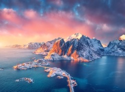 Beautiful landscape with blue sea, snowy mountains, rocks and islands, village, rorbu, road, bridge and pink sky at sunrise. Aerial view. Hamnoy in snow in winter in Lofoten islands, Norway. Top view