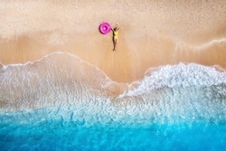 Aerial view of the lying beautiful young woman with pink swim ring on the sandy beach near sea with waves at sunset. Summer vacation in Lefkada island, Greece. Top view of slim girl, clear azure water