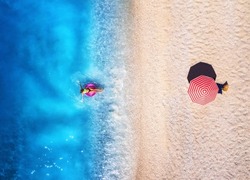 Aerial view of a woman swimming with pink swim ring in blue sea, sandy beach and red umbrella at sunset in summer. Tropical landscape with girl, clear water, waves. Top view. Lefkada island, Greece