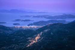 Small village on the mountain and city on the sea cost at night. Aerial view. Landscape with city lights, port, town, buildings, forest on hills, islands, blue water and purple sky at dusk. Greece