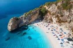 Aerial view of blue sea, rock, sandy beach with umbrellas at sunrise in summer. Porto Katsiki, Lefkada island, Greece. Beautiful landscape with sea coast, swimming people, trees, azure water. Top view