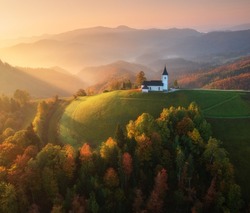 Aerial view of small church in fog and colorful forest at sunrise in autumn. Slovenia. Top view of beautiful chapel on mountain in mist, green meadows, trees, orange sky at dawn in fall. Landscape 