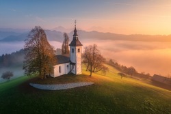 Aerial view of small church on the hill and orange low clouds at sunrise in autumn. Slovenia. Top view of beautiful chapel on mountain in fog, green meadows, trees, sky at dawn in fall. Landscape