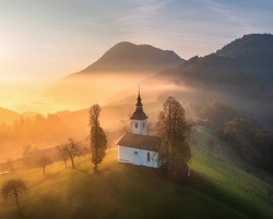 Aerial view of small church on the hill in low clouds at golden sunrise in autumn. Slovenia. Top view of beautiful chapel, mountain village, fog, green meadows, trees, sky at dawn in fall. Landscape 