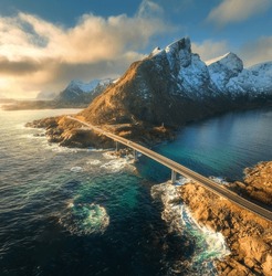 Aerial view of bridge, sea with waves and mountains at sunset in Lofoten Islands, Norway. Landscape with beautiful road, water, rocks, blue sky with clouds and golden sunlight. Top view from drone