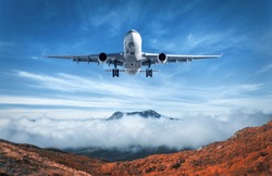 Airplane is flying over low clouds and mountains with autumn forest. Amazing landscape with passenger airplane, trees, mountains, blue cloudy sky. Passenger aircraft. Business travel. Commercial plane