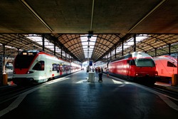Perspective view of a platform in Lucerne Central Station with sunlight cast on trains parking by the platform & passengers hurrying for boarding ~ A beautiful corner of the railway terminal in Lucern