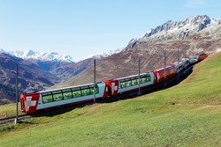 Tourists riding a Glacier Express on a brisk fall day & enjoying the view thru panoramic windows of grassy meadows on the hillside & snowy mountains under blue clear sky in Andermatt, Uri, Switzerland