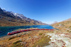 A local train of Rhaetian railway traveling on the lakeside meadows by Lago Bianco and the alpine mountains towering under blue clear sky on a sunny autumn day, in Bernina Pass, Grisons, Switzerland