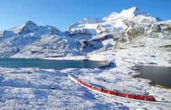 Aerial view of a local train of Rhaetian Railway (RhB) traveling by the lake Lago Bianco, with alpine mountains covered by snow under blue sunny sky, in Bernina Pass, canton of Grisons, Switzerland