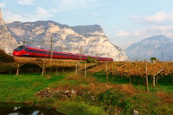 Beautiful scenery of a fast train traveling through the vineyard field on a sunny autumn day, with rugged cliffs of alpine mountains dominating the background, in Mezzocorona, Trentino, Italy, Europe