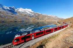 A train of Rhaetian Railway travels along Lago Bianco in Bernina Pass on a brisk autumn day with mountain peaks covered by snow and reflected in the water under blue clear sky, in Grisons, Switzerland