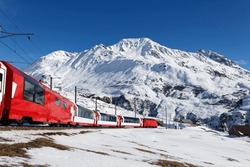 On a sunny winter day, tourists ride in a Glacier Express train and, thru the panoramic windows, enjoy the view of snowy Rossbodenstock mountain under blue clear sky, in Andermatt, Uri, Switzerland