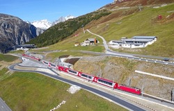 Aerial view of a Glacier Express train departing from Natschen Station, with a winding highway on grassy meadows and rugged snowy mountains under blue sunny sky, in Andermatt, Uri, Switzerland