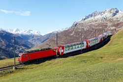 Tourists ride in a Glacier Express on Matterhorn Gotthard Railway on a brisk fall day and, thru panoramic windows, enjoy the view of snowy mountains under blue clear sky in Andermatt, Uri, Switzerland