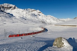 A train of RhB (Rhaetian Railway) travels by the lake Lago Bianco with alpine mountains covered by snow under blue clear sky on a sunny winter day, in Bernina Pass, canton of Graubünden, Switzerland
