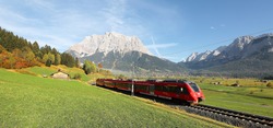 View of a fast train traveling through green fields with magnificent Mountain Zugspitze in the background on a crisp sunny day in Lermoos, Tirol, Austria ~ Brisk autumn scenery of Tyrolean countryside