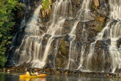 Artificial waterfall above river in summer day. Water cascade flowing on stones. Splash stream between the trees. Cold current descends on the shore. People in yellow kayak look at beautiful waterfall