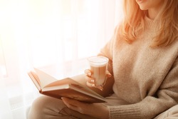 Young beautiful woman near window beige knitted sweater read book, wish list, daily planner, notepad, goals. Relax concept. Hold cappuccino glass of coffee with white foam. Sun's rays shine into room