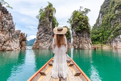 Back View of Young Female Tourist in Dress and Hat at Longtail Boat near Three rocks with Limestone Cliffs at Cheow Lan Lake. Woman on Boat in Ratchaprapha Dam Khao Sok National Park in Thailand