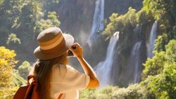 Millennial traveller or Inspiring travel blogger make photos for social media on beautiful tropical waterfall, amazing nature. Wanderlust traveling. Female traveler in trip in Southeast Asia