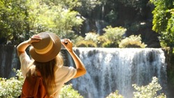 Happy female tourist in trip, journey. Young woman blogger, zoomer traveler make photo for social media of amazing waterfall, nature on trendy retro camera. Wanderlust, travel concept. Cinematic shot.
