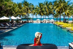 Luxury beach vacation in tropical beach hotel. Tourist woman in red dress relax near blue swimming pool in modern resort. Female traveler on sea vacation.