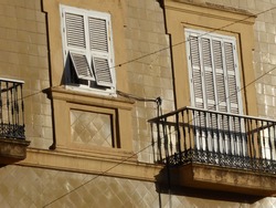 Old windows in old Mediterranean style architecture. A pretty cast iron balcony with its silhouette from bright sunshine. Two shutters are open on a slant. The wall is unusually tiles in two patterns