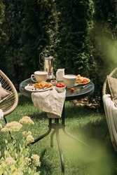 The summer breakfast with cups of coffee, croissants and strawberry on the small round table and chairs nearby in the green garden, visible from behind the growing flowers in a sunny day.