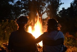 Traditional midsummer celebration in Latvia. Man and woman sitting,  at a big bonfire, holding hands, looking into bright flames. Wearing flower wreaths. 