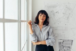 Female architect standing in office beside architecture drawings. Woman entrepreneur standing in office making presentation.