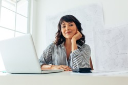 Smiling female architect working on laptop in office. Entrepreneur sitting at workplace with architecture drawings in the background.
