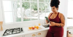 Wide angle shot of beautiful woman looking at cell phone while standing in kitchen. Healthy female in fitness wear watching social media video on mobile phone at home.