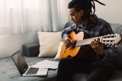 Black male musician learning guitar lessons online. Young guitarist holding a guitar while watching a music tutor on his laptop. Man studying music at home during quarantine.