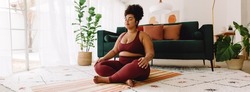 Plus size woman sitting cross legged and closed eyes in living room. Pretty female in fitness wear practicing meditation at home.