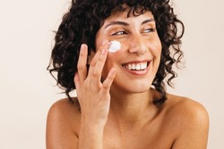 Happy young woman smiling cheerfully while applying moisturising cream on her face. Attractive young woman treating her facial skin with a nourishing beauty product.