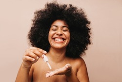 Gorgeous young woman smiling cheerfully while dropping cosmetic face serum onto her palm. Happy woman with Afro hair treating her skin with a moisturizing beauty product.