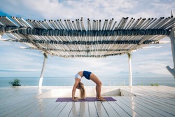 Woman with long hair and slim body make morning yoga exercise on the yoga mat on white wooden terrace. Yoga pose. Bridge pose. 
