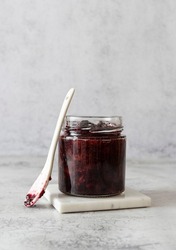 A glass jar with blackberry jam on a white marble stand. A white ceramic spoon next to the jar. Light background