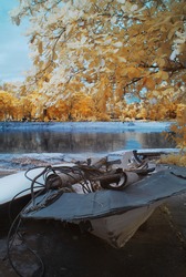 An Infrared Photography view of an abandoned boats by a lakeside surrounded by golden foliage trees.