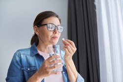 Mature middle-aged woman in casual clothes at home holding pill and glass of fresh water. Headaches, depression, medicine supplements vitamins, sedatives, menopause