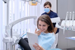 Happy middle aged woman with doctor dentist looking in mirror at teeth, sitting in dental chair. Medicine, dentistry and health care concept