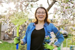Spring gardening, portrait of mature smiling woman with rake looking at camera. Blooming trees in the garden background, seasonal cleaning in the garden, hobbies and leisure