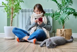 Woman at home using smartphone, warming with cat near heating radiator