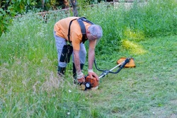A mature man in protective clothing, and gloves with a trimmer. Mowing tall grass and the weeds with a trimmer. Selectively focus