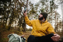 A bearded man rests on a fallen tree in the middle of the forest. A man uses a mobile phone to search GPS.