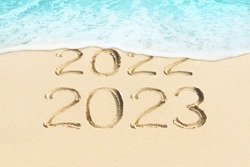New Year concept photo. Text 2023 and 2022 handwritten in sand surface. Blue ocean wave washing away numbers on beach. 